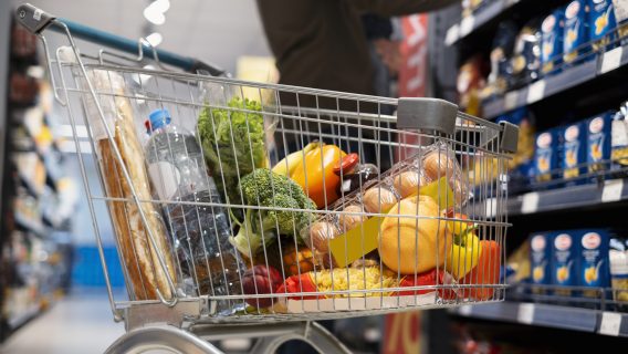 Image shows a shopping trolley containing food and a person selecting items from a shelf.